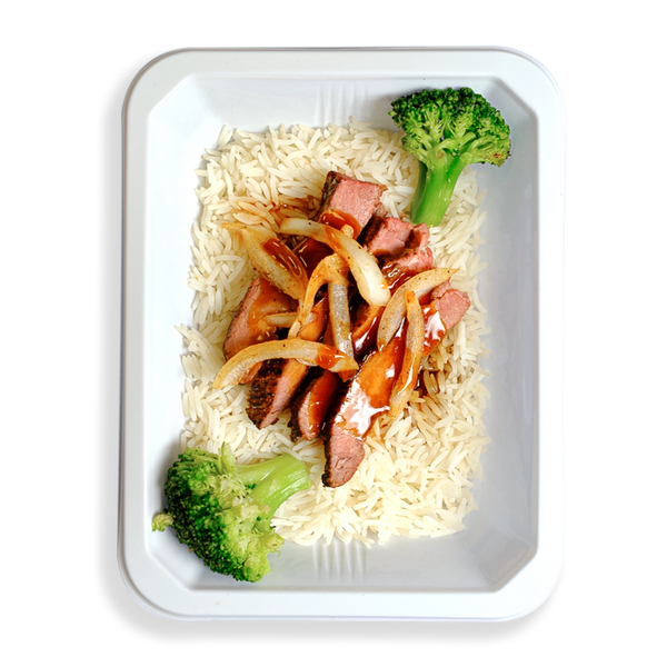 Thinly sliced flank steak over white rice, drizzled with low-sodium teriyaki sauce, is neatly arranged in a white to-go container. Bright green broccoli and julienned onions add a fresh, vibrant touch to this thoughtfully prepared meal—ideal for those who value healthy eating without compromising on flavor or quality.