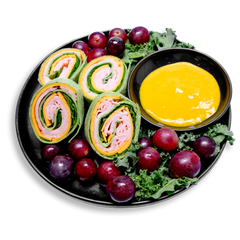A black plate filled with vibrant health food: spinach tortilla pinwheels tightly rolled with nitrate-free smoked turkey breast, cheddar cheese, and fresh spinach leaves. Surrounding the wraps are plump red grapes and a bed of curly kale, with a side of golden sweet mustard dipping sauce in a black ramekin. This healthy prepared meal balances savory and sweet for a visually striking and nutritious snack or lunch option.