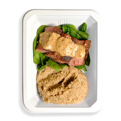 Sliced flank steak topped with creamy peppercorn sauce rests on a bed of fresh spinach, alongside a smooth, herbed white bean purée—all neatly arranged in a white to-go container. The presentation suggests it's made for those with full schedules who still prioritize eating well, even when cooking isn't an option.